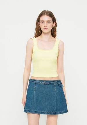 Jeune femme aux cheveux ondulés portant un haut jaune pâle sans manches avec des bords festonnés et une jupe en jean bleue avec une ceinture.