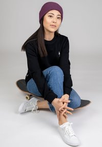 Zwarte langslavige shirt met een wit boomblogo, blauwe denim jeans en witte sneakers; model draagt een paarse geribde beanie, zittend op een skateboard.
