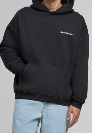 Person trägt einen schwarzen Hoodie mit dem Schriftzug "Slow Sunday Club" auf der linken Brustseite, hellblaue Jeans und hat eine Hand in der Kängurutasche.