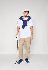 Man wearing white cap, white polo, navy sweater over shoulders, beige pants, blue shoes, and golf glove, holding golf club standing against plain background.