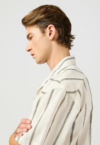 Camicia colletto in crema con righe verticali grigie, realizzata in tessuto leggero. Presenta maniche corte e un design a vestibilità rilassata.