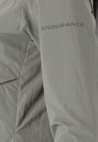 Ljusgrå jacka med en mjuk textur, lätt glansig yta och "ENDURANCE"-logotyp i kontrasterande grå på vänster ärm.