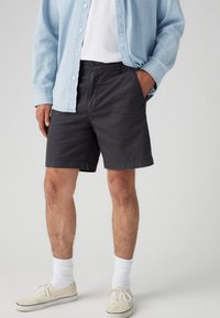 Pantaloni scurți gri închis cu croială dreaptă, buzunare laterale și bucle pentru curea, purtați cu o cămașă butonată de culoare albastru deschis și adidași albi.