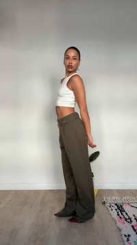 Žena v bielom krátkom tielku a voľných olivových nohaviciach stojaca zboku v minimalistickej miestnosti, kde je vidieť rastlinu a vzorovaný koberec.