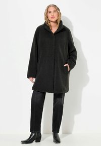 Cappotto nero, oversize, in pelliccia sintetica con colletto e tasche. Il modello indossa jeans neri e stivaletti. Tessuto liscio con una vestibilità rilassata.