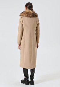 Cappotto di lana beige con un grande colletto in pelliccia marrone, taglio dritto, maniche lunghe e cucitura sul retro dettagliata, abbinato a stivali in pelle neri al ginocchio.