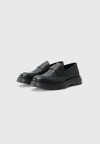Black leather slip-on shoes with a thick, textured rubber sole and a raised band at the front featuring embossed logo detailing.