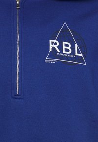 Felpa blu con zip realizzata in tessuto morbido, dotata di un logo triangolare bianco con la scritta "RBL" e le coordinate, e una zip argentata.
