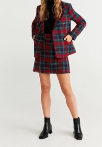 Ensemble veste et jupe en tissu texturé à carreaux rouges et bleu marine, avec poches plaquées et devant croisé. Associé à des bottines noires.