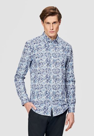 Młody mężczyzna ubrany w niebiesko-białą koszulę w kwiaty z długim rękawem i czarne spodnie, stojący z jedną ręką w kieszeni.