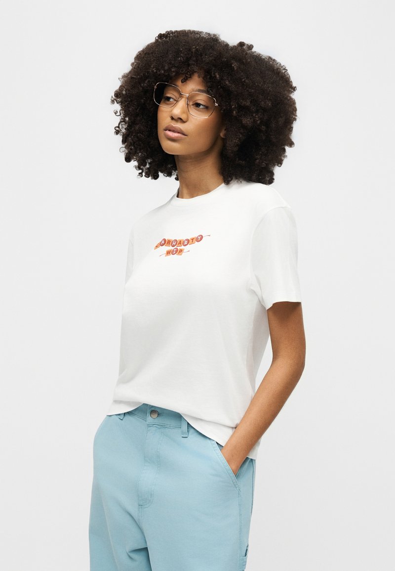 Femme aux cheveux bouclés et portant des lunettes, vêtue d'un T-shirt blanc graphique et d'un pantalon bleu clair, debout avec une main dans la poche, regardant sur le côté.