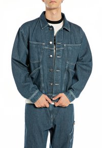 Denim jacket in medium blue, featuring a button-up front, large patch pockets, and a relaxed fit with a slightly structured design.