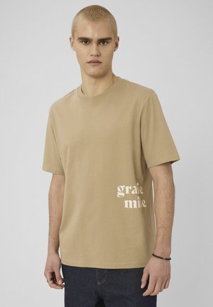 Jeune homme aux cheveux blonds courts portant un t-shirt beige avec un texte blanc sur le côté et un jean foncé, sur un fond uni.
