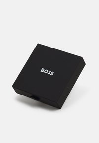 Boîte en carton noir à texture lisse, avec le mot "BOSS" en argent sur le dessus et un petit onglet pour l'ouvrir.