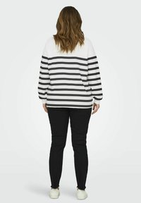 Camisa de manga larga a rayas blancas y negras con un ajuste relajado, combinada con pantalones negros y zapatillas blancas, vista desde la parte posterior.