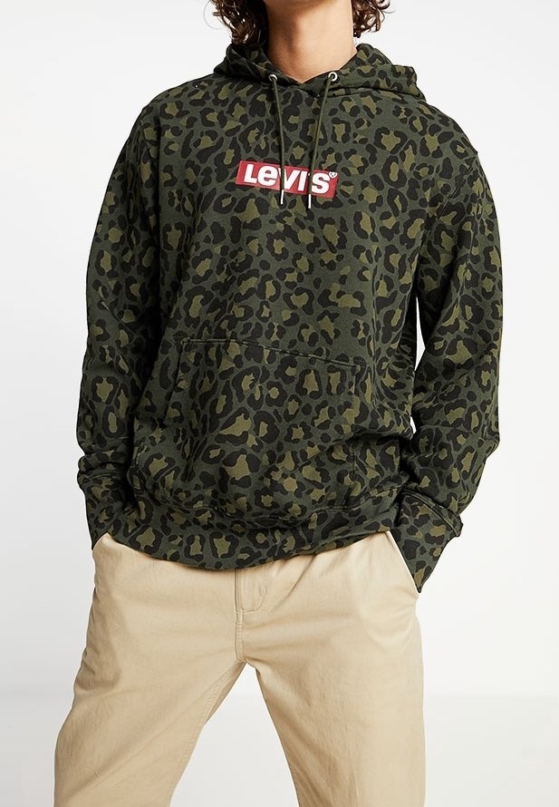 Personne portant un sweat à capuche Levi’s vert et noir à motif léopard avec poche avant et pantalon beige, mains partiellement dans les poches.