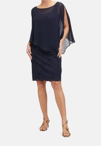 Femme portant un haut à superposition en chiffon bleu marine avec des manches ouvertes, une jupe en dentelle bleu marine longueur genou, et des sandales à talons hauts dorées à brides.