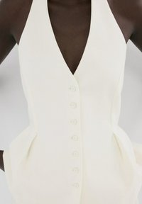 Mujer con vestido blanco sin mangas con botones, escote en V profundo y cintura ajustada, de pie con las manos en las caderas.