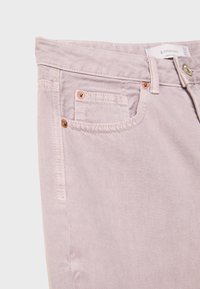 Jean en denim rose clair en gros plan montrant la poche avant, la petite poche, les passants de ceinture et les rivets en cuivre sur fond uni.
