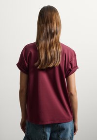 T-shirt bordeaux con maniche corte a risvolto, tessuto morbido, vestibilità rilassata e scollatura rotonda, indossato con pantaloni shorts in denim.