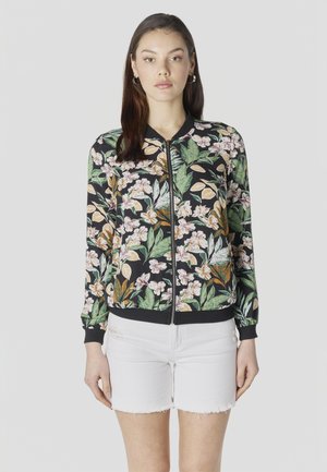 Mujer con cabello largo y oscuro, vestida con una chaqueta negra con estampado floral y cremallera, y pantalones cortos blancos con dobladillo deshilachado, de pie frente a un fondo liso.