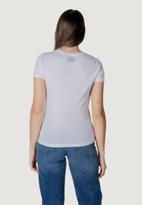 T-shirt bianca in cotone con maniche corte, design fitted, con un piccolo logo EA7 viola sulla parte superiore della schiena. Texture liscia.