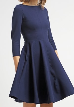 Vrouw draagt een donkerblauwe jurk tot op de knie met driekwart mouwen en een nauwsluitend lijfje dat uitloopt in een volle rok.