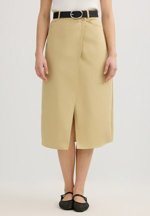 Falda beige a la rodilla con abertura frontal, combinada con cinturón negro y zapatos planos negros en persona de pie.