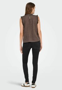 Žena v hnědé bezrukávové blůze s volánkovými okraji a černých skinny džínách, viděná zezadu, stojící na bílém pozadí.