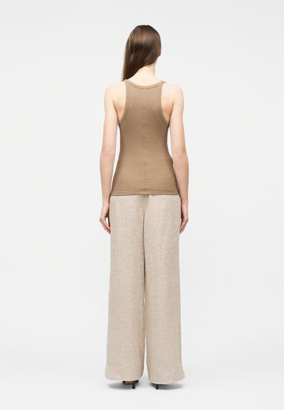 Femme debout de dos portant un haut marron sans manches ajusté et un pantalon beige à jambes larges avec des talons noirs sur un fond blanc uni.