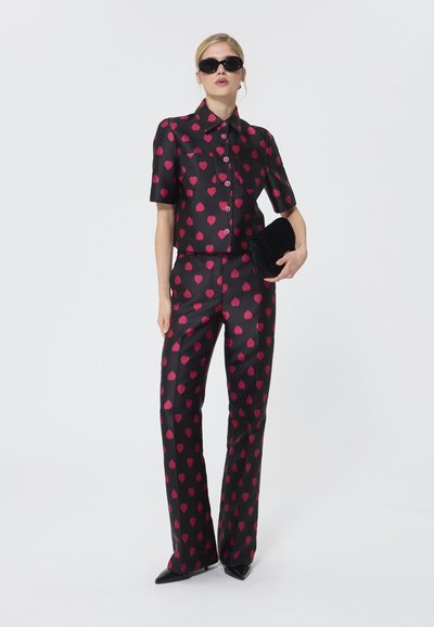 Schwarzes Outfit bestehend aus einem kurzärmeligen Shirt und weit geschnittenen Hosen mit einem roten Herzmuster. Ergänzt durch schwarze Sonnenbrille und eine Clutch.