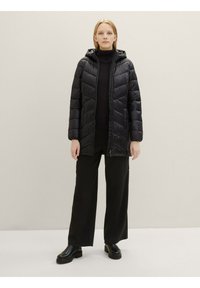 Giubbotto invernale nero con cappuccio, caratterizzato da un design trapuntato, chiusura frontale con zip e maniche lunghe. Indossato sopra un dolcevita nero e pantaloni.
