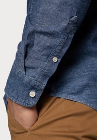 Mężczyzna w niebieskiej, dżinsowej koszuli z długim rękawem i zapinanymi na guziki mankietami oraz beżowych spodniach, z ręką włożoną do kieszeni.