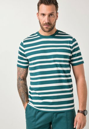 Mand med kort mørkt hår og skæg iført en kortærmet skjorte med teal og hvide striber og brystlomme samt teal bukser, stående foran en hvid baggrund.