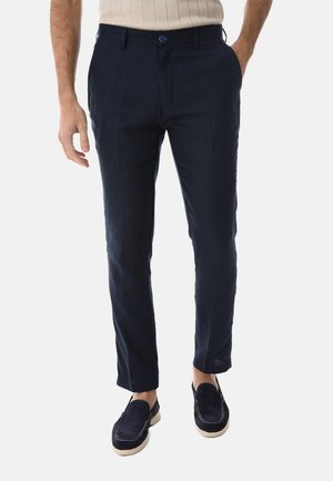 Uomo che indossa pantaloni aderenti blu navy, camicia beige a maniche corte e scarpe slip-on blu navy con suole bianche, mani nelle tasche.