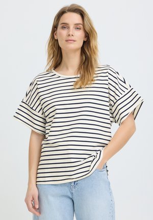 Femme aux longs cheveux blonds portant un t-shirt à manches courtes blanc à rayures bleu marine et un jean bleu clair, main dans la poche, fond neutre.