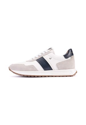 Zapatilla blanca y beige con franja negra, diseño con cordones y suela de goma marrón, mostrada de lado sobre un fondo blanco.