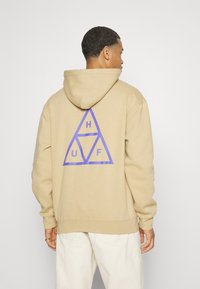 Sudadera beige con un gran logo de triángulo geométrico morado en la parte posterior. Fabricada con material suave, tiene un corte relajado y capucha.