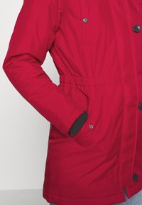 Manteau d'hiver rouge avec taille élastique, boutons-pression et poignet noir visible à l'intérieur de la poche tenu par une personne.