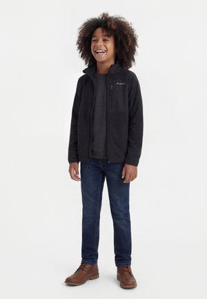 Hymyilevä lapsi kiharat hiukset, musta vetoketjullinen takki, tumma harmaa paita, siniset farkut ja ruskeat nauhalliset saappaat päällään, seisoo valkoisella taustalla.