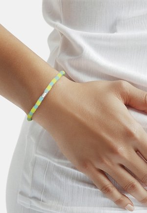Wrist with a beaded bracelet in alternating pastel yellow and blue, featuring a small silver rectangular charm, worn over white textured fabric.