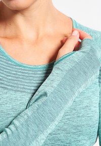 Długi rękaw, niebiesko-zielony top sportowy z teksturowanej tkaniny, z prążkowanym wzorem wokół dekoltu oraz otworami na kciuki na rękawach.