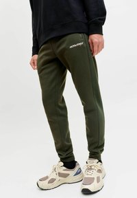 Olivengrønne joggebukser med ribbede sidelæg, elastisk talje og broderet logo. Kombineret med beige træningssko og sort sweater.