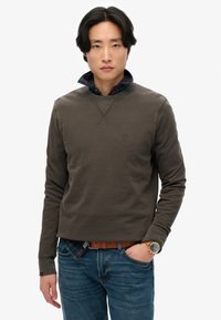 Sudadera verde oliva con cuello redondo y logo en el pecho, superpuesta a una camisa de cuadros. Combinada con jeans azules y un reloj de pulsera, exhibiendo un estilo casual.