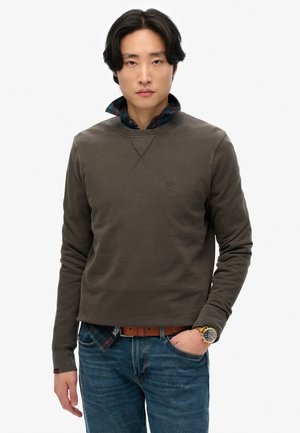 Olijfgroene sweatshirt met ronde hals en logoprint op de borst, gedragen over een geruit shirt. Gecombineerd met blauwe jeans en een horloge, wat een casual stijl laat zien.