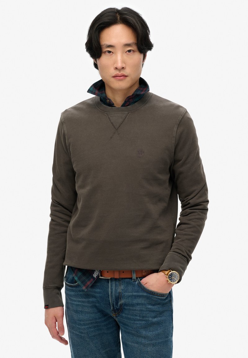Sudadera verde oliva con cuello redondo y logo en el pecho, superpuesta a una camisa de cuadros. Combinada con jeans azules y un reloj de pulsera, exhibiendo un estilo casual.
