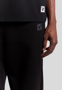 Schwarzes Baumwoll-T-Shirt und Hose mit figurbetontem Schnitt. Verfügt über kleine Logo-Patches auf dem T-Shirt und der Hose in kontrastierenden Farben.