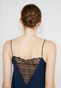 Marinblå topp med tunna axelband och en djup spets V-rygg med scallopade kanter och intrikata blomster mönster. Slät tygtextur.