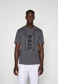 Pilkas trumpas rankovėmis marškin�ėliai su ryškiu juodu "BOSS" logotipu priekyje ir tiesiu pjaustymu. Modelis vilki baltas kelnes.