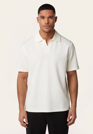 Mann mit kurzen Haaren, der ein weißes Polohemd mit kurzen Ärmeln und schwarze Hosen trägt, vor einem schlichten, hellen Hintergrund stehend.